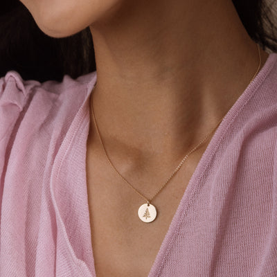 A close-up of a model wearing a delicate gold necklace with a small round pendant featuring a pine tree design, styled with a soft pink top.