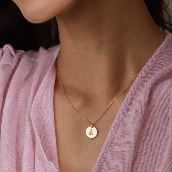 A close-up of a model wearing a delicate gold necklace with a small round pendant featuring a pine tree design, styled with a soft pink top.