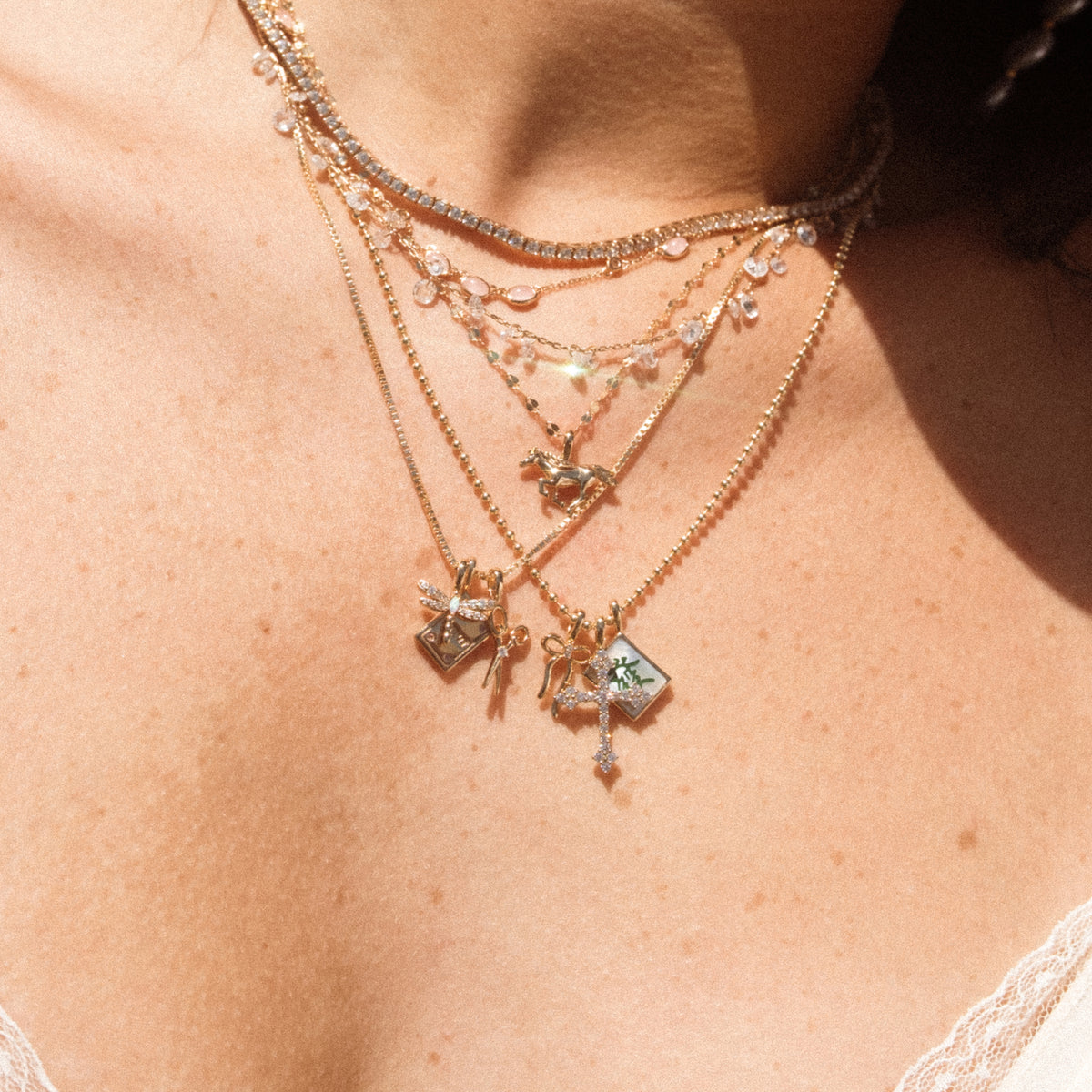 A close up lifestyle shot of a person's neck wearing multiple layered gold charm necklaces in varying lengths. Across the different chain styles, some of the charms featured are a dragonfly, a cross with clear stones, a bow, and a running horse.