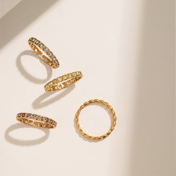 Four gold rings, each with a different color of square bezel set gemstones side by side creating the band, set against a white background with light shadows.