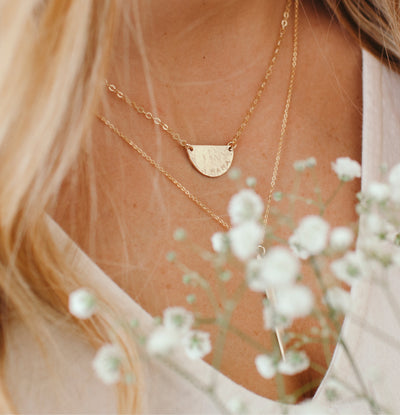 A close-up lifestyle shot of a woman wearing a delicate gold necklace with a textured, semi-circular disc pendant layered over a thin chain necklace. The image is styled with soft-focus white baby's breath flowers in the foreground and features warm, natural lighting.