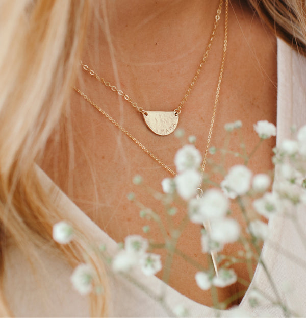 A close-up lifestyle shot of a woman wearing a delicate gold necklace with a textured, semi-circular disc pendant layered over a thin chain necklace. The image is styled with soft-focus white baby's breath flowers in the foreground and features warm, natural lighting.