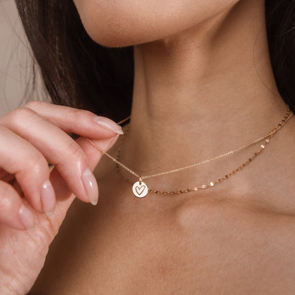 A close-up of a model wearing two delicate layered gold necklaces, one with a small heart-shaped pendant, while her hand gently touches the chain.