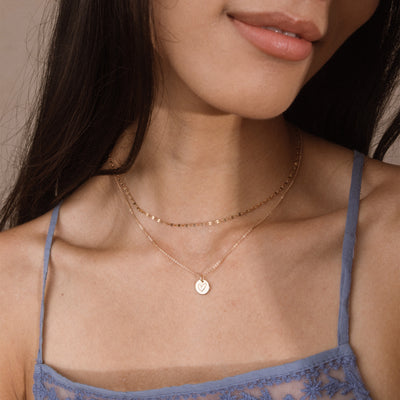 A close-up of a model wearing two delicate layered necklaces: one gold chain with circular disc links and one gold chain with a small round pendant engraved with a heart, paired with a blue lace camisole.