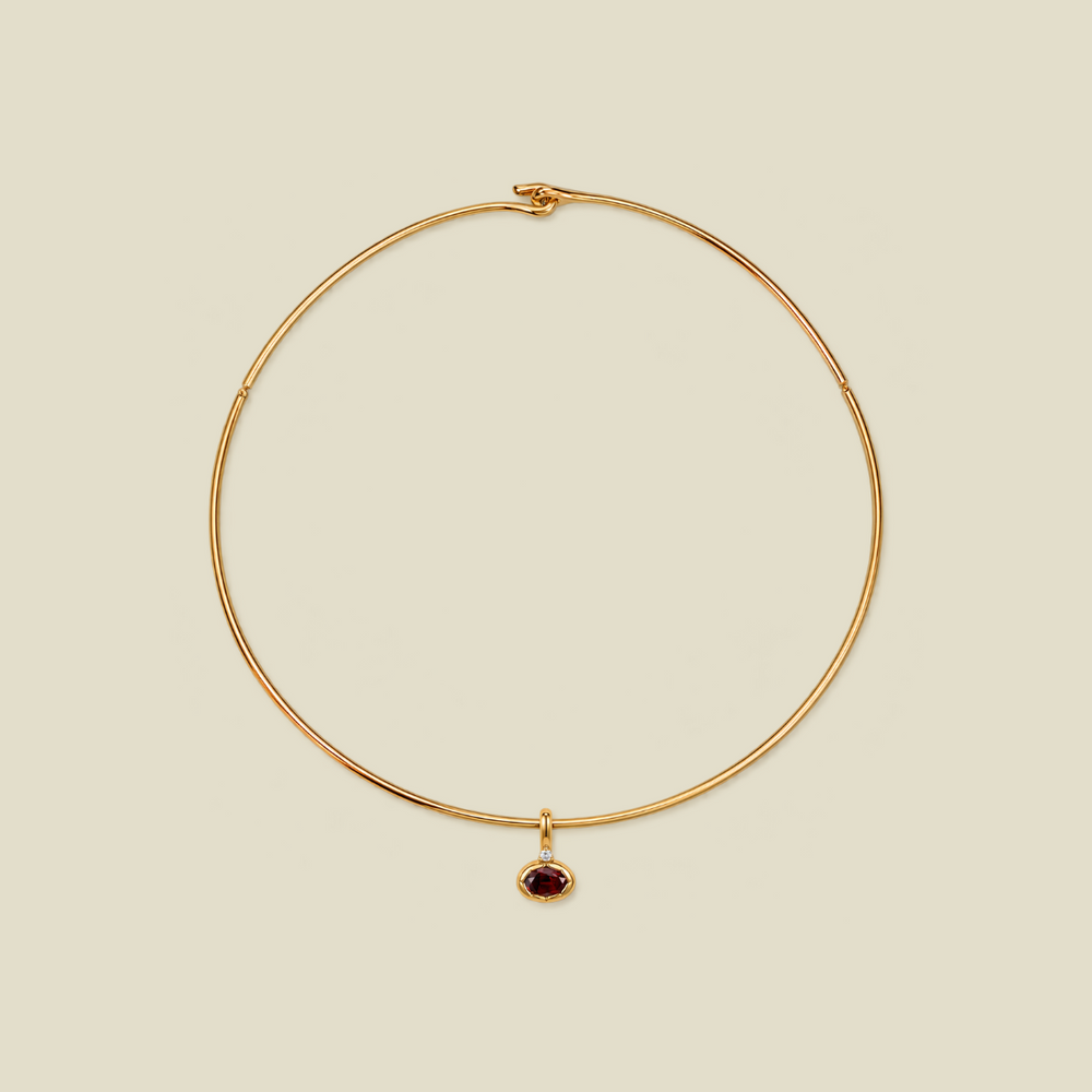 A delicate gold wire circle necklace with a small oval dark red gemstone pendant, displayed on a solid light beige background.