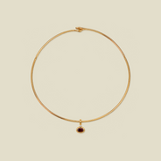 A delicate gold wire circle necklace with a small oval dark red gemstone pendant, displayed on a solid light beige background.
