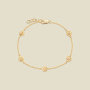 A delicate gold chain bracelet with five small, textured carnation charms spaced evenly along the chain, displayed on a solid light beige background.