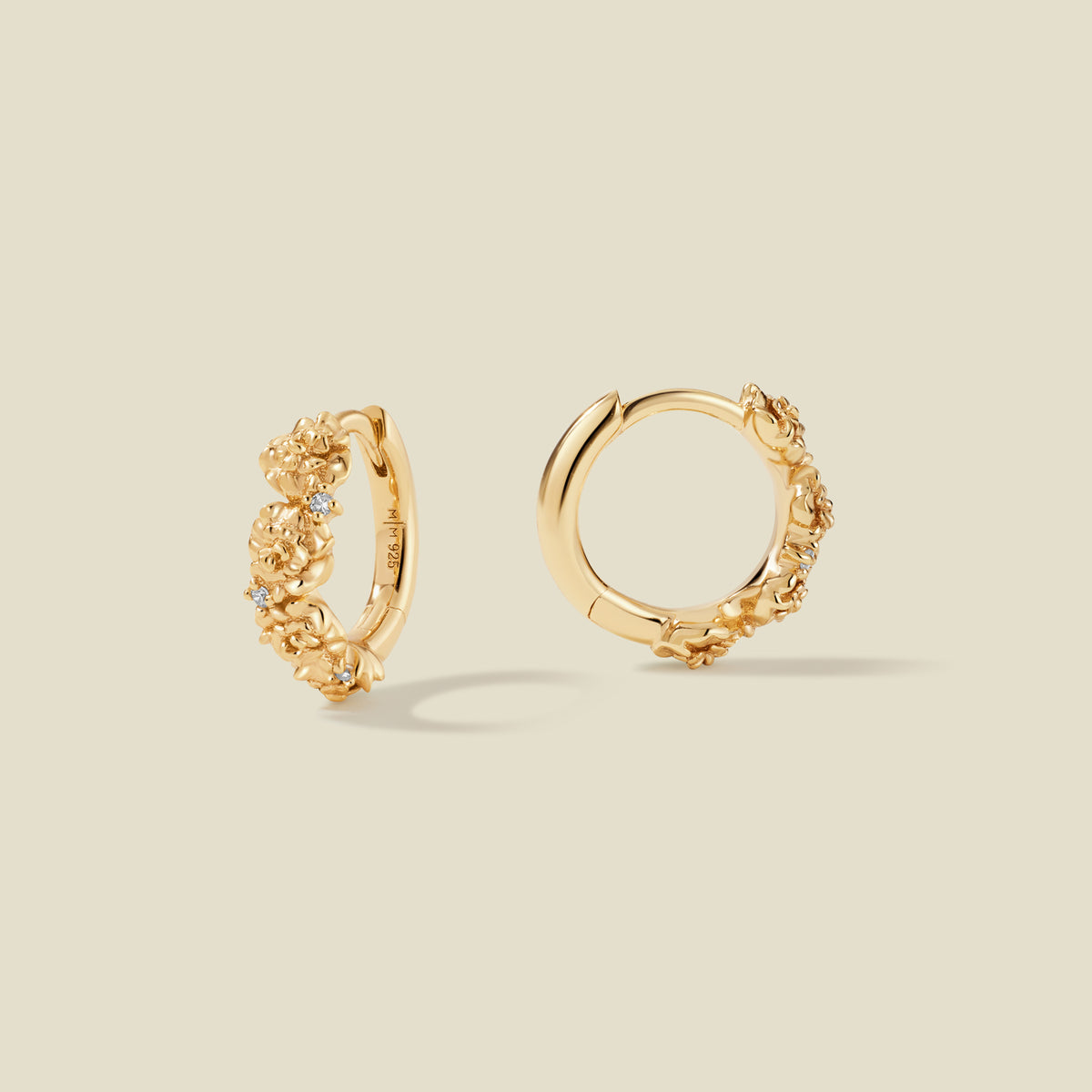 A pair of gold hoop earrings adorned with carnation flowers stacked along the hoop, with scattered small clear accent stones, set against a solid beige background.