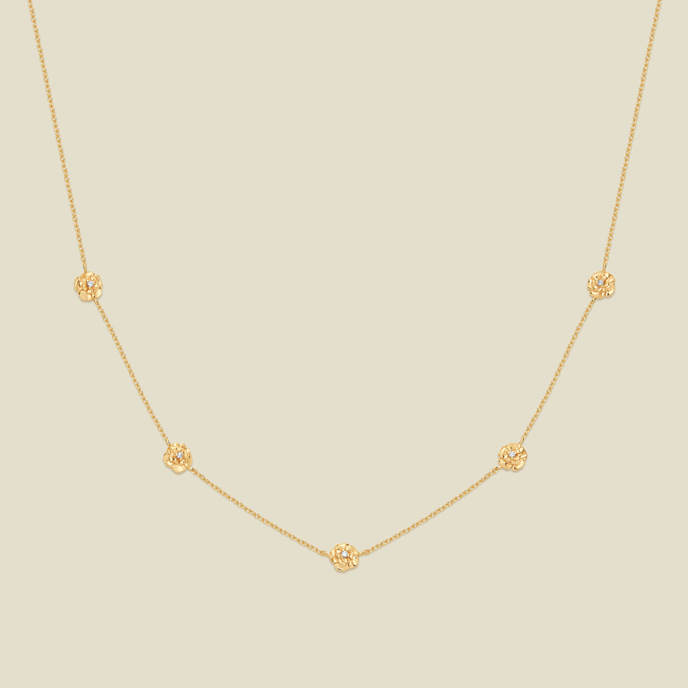 A delicate gold chain necklace featuring five small, evenly spaced textured carnation flower motifs set with tiny clear stones, displayed on a solid light beige background.