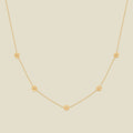A delicate gold chain necklace featuring five small, evenly spaced textured carnation flower motifs set with tiny clear stones, displayed on a solid light beige background.