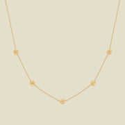 A delicate gold chain necklace featuring five small, evenly spaced textured carnation flower motifs set with tiny clear stones, displayed on a solid light beige background.