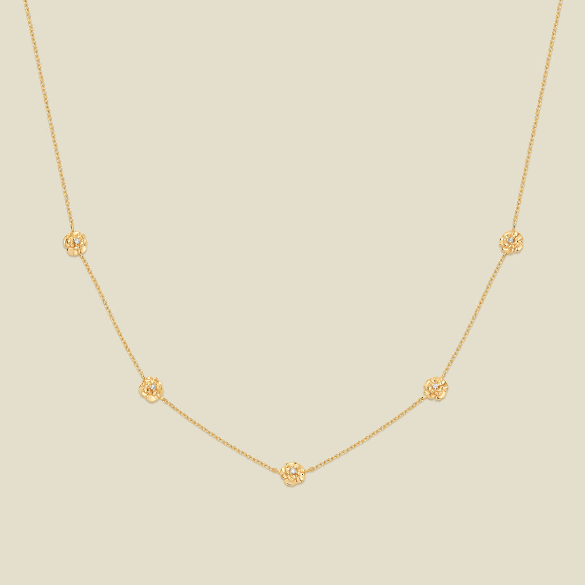 A delicate gold chain necklace featuring five small, evenly spaced textured carnation flower motifs set with tiny clear stones, displayed on a solid light beige background.