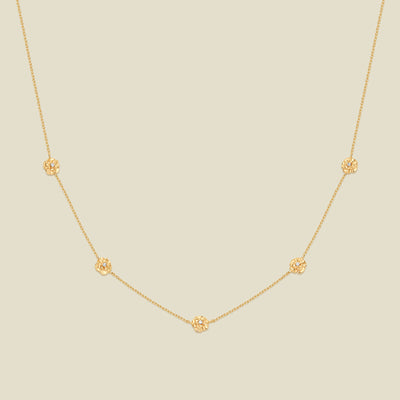 A delicate gold chain necklace featuring five small, evenly spaced textured carnation flower motifs set with tiny clear stones, displayed on a solid light beige background.