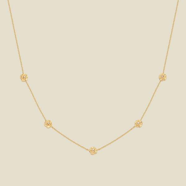 A delicate gold chain necklace featuring five small, evenly spaced textured carnation flower motifs set with tiny clear stones, displayed on a solid light beige background.