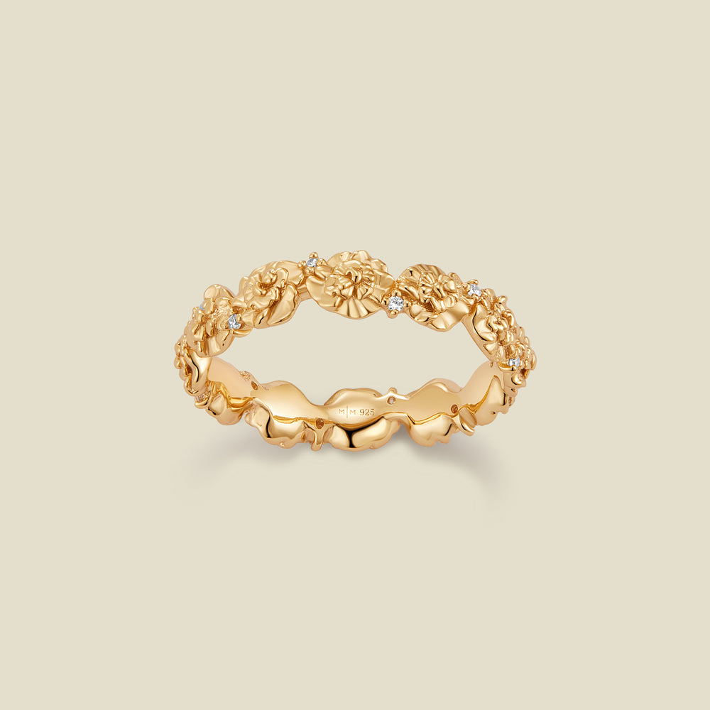 A gold floral-etched ring band with textured carnation flowers and small clear gemstones, shown on a plain warm beige background. 