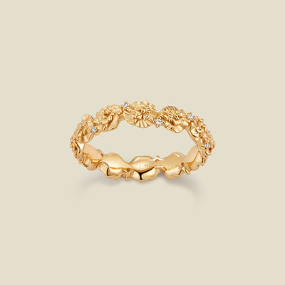 A gold floral-etched ring band with textured carnation flowers and small clear gemstones, shown on a plain warm beige background. 