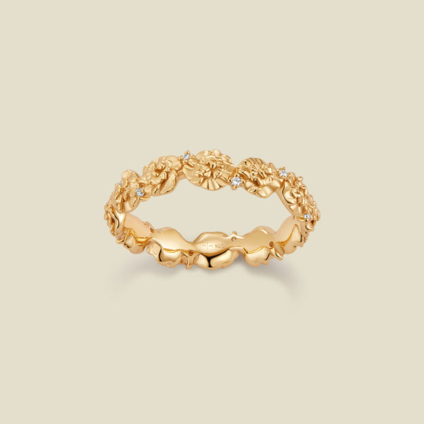 A gold floral-etched ring band with textured carnation flowers and small clear gemstones, shown on a plain warm beige background. 