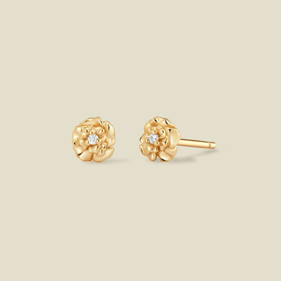 A pair of gold carnation flower stud earrings, each featuring a small round clear gemstone at the center, displayed on a solid light beige background.