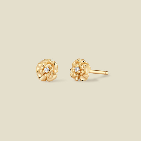 A pair of gold carnation flower stud earrings, each featuring a small round clear gemstone at the center, displayed on a solid light beige background.