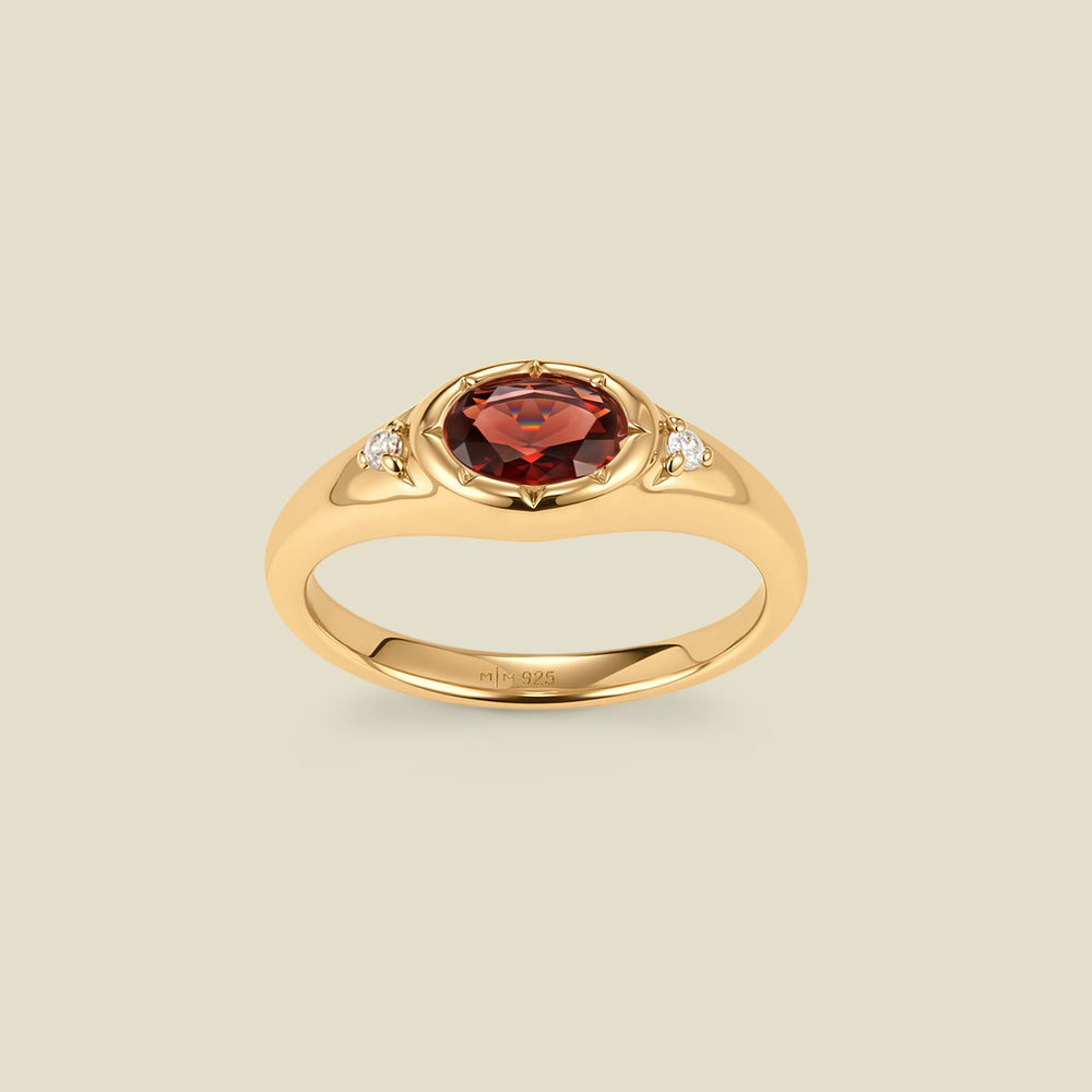 A gold signet ring with a red center oval bezel-set gemstone, and two small clear accent stones on either side, set against a solid beige background.