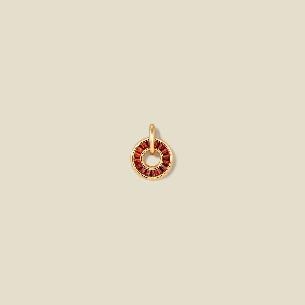 A circular gold pendant featuring red baguette-cut stones around a hollow center, displayed against a solid light beige background.