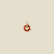 A circular gold pendant featuring red baguette-cut stones around a hollow center, displayed against a solid light beige background.