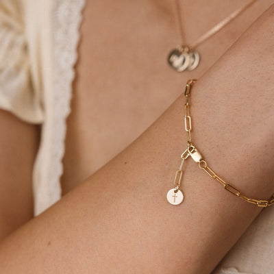 A close-up lifestyle shot of a delicate gold paperclip chain bracelet with a small round disc charm engraved with a cross, worn on a person's wrist.