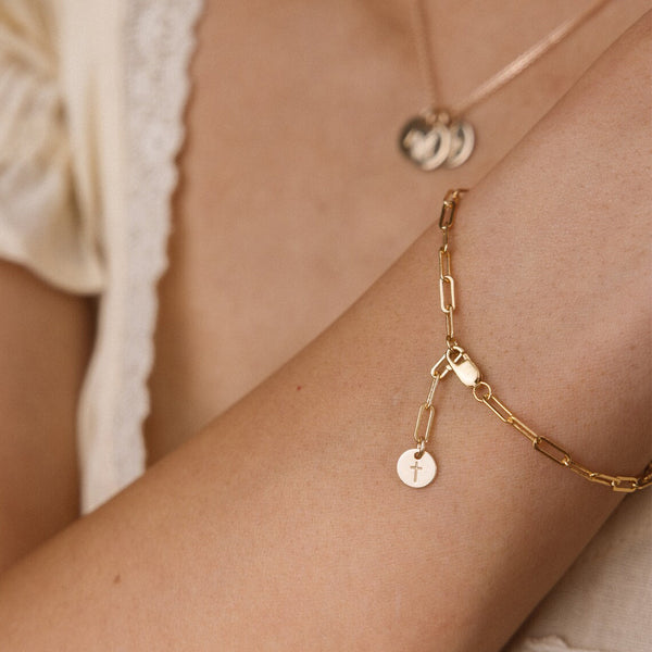 A close-up lifestyle shot of a delicate gold paperclip chain bracelet with a small round disc charm engraved with a cross, worn on a person's wrist.