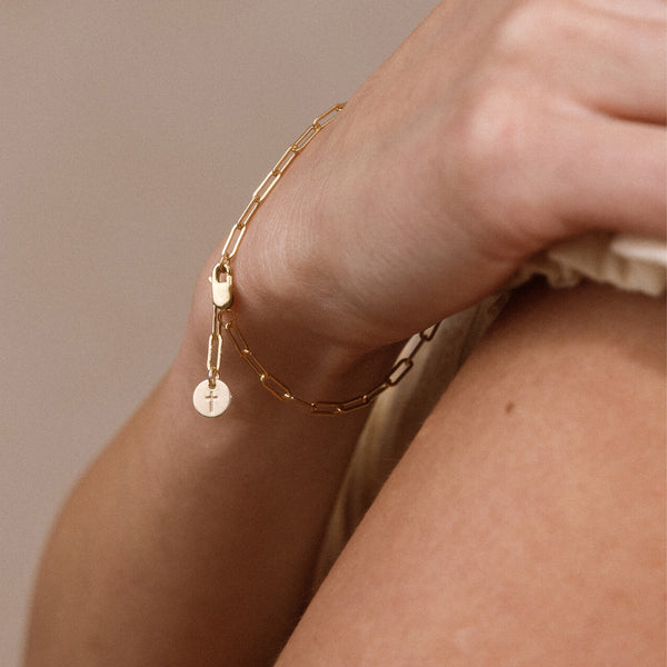A delicate gold chain bracelet with a small round charm engraved with a cross, worn on a person's wrist against a soft beige background.