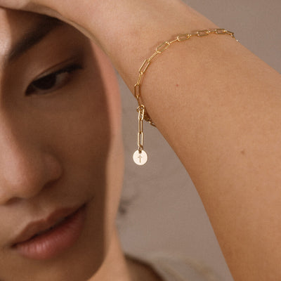 A close-up lifestyle shot of a model's forearm wearing a delicate gold chain bracelet with a small circular disc charm, engraved with a cross.