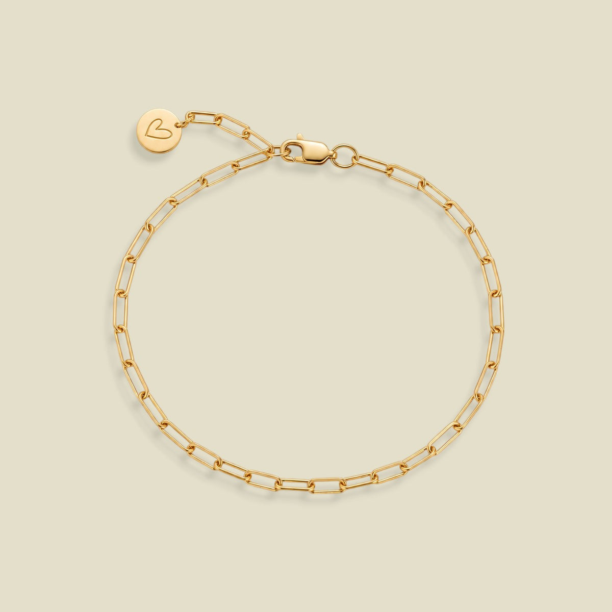 A gold paperclip chain bracelet with a lobster clasp and a small round engraved charm with a heart on the end of the extender, displayed on a light beige background.