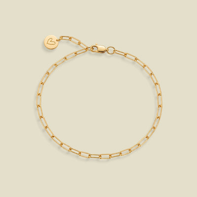 A gold paperclip chain bracelet with a lobster clasp and a small round engraved charm with a heart on the end of the extender, displayed on a light beige background.
