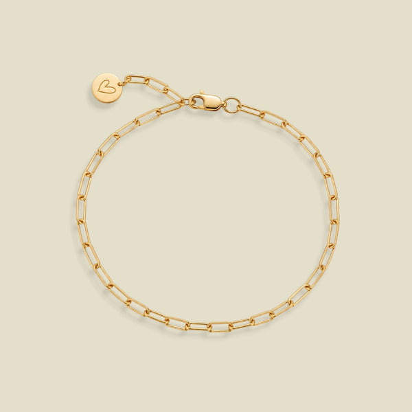 A gold paperclip chain bracelet with a lobster clasp and a small round engraved charm with a heart on the end of the extender, displayed on a light beige background.