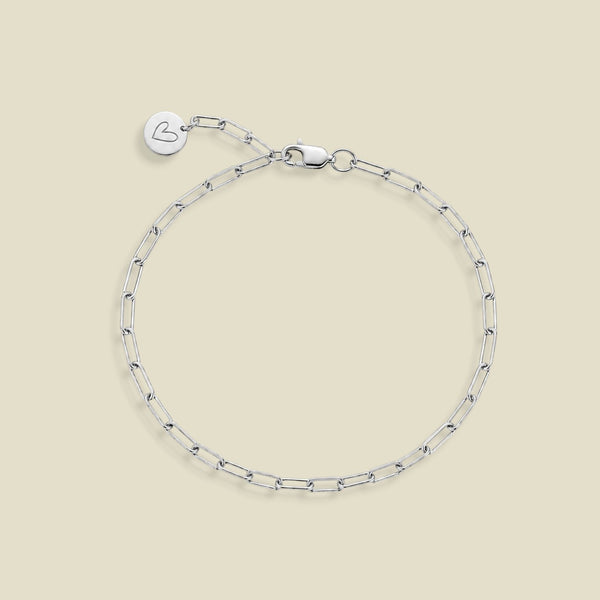 A silver paperclip chain bracelet with a lobster clasp, an adjustable extension chain, and a small round engraved charm with a heart on the end of the extender, displayed on a solid beige background.