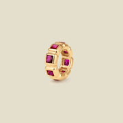 A gold ring charm with multiple bezel set square-cut deep red gemstones set side by side to create the band, shown on a solid beige background.