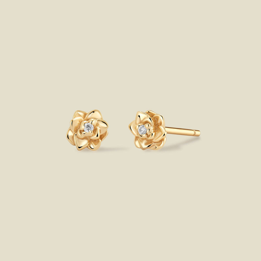 A pair of gold lotus flower stud earrings, each featuring a small round clear gemstone at the center, displayed on a solid light beige background.