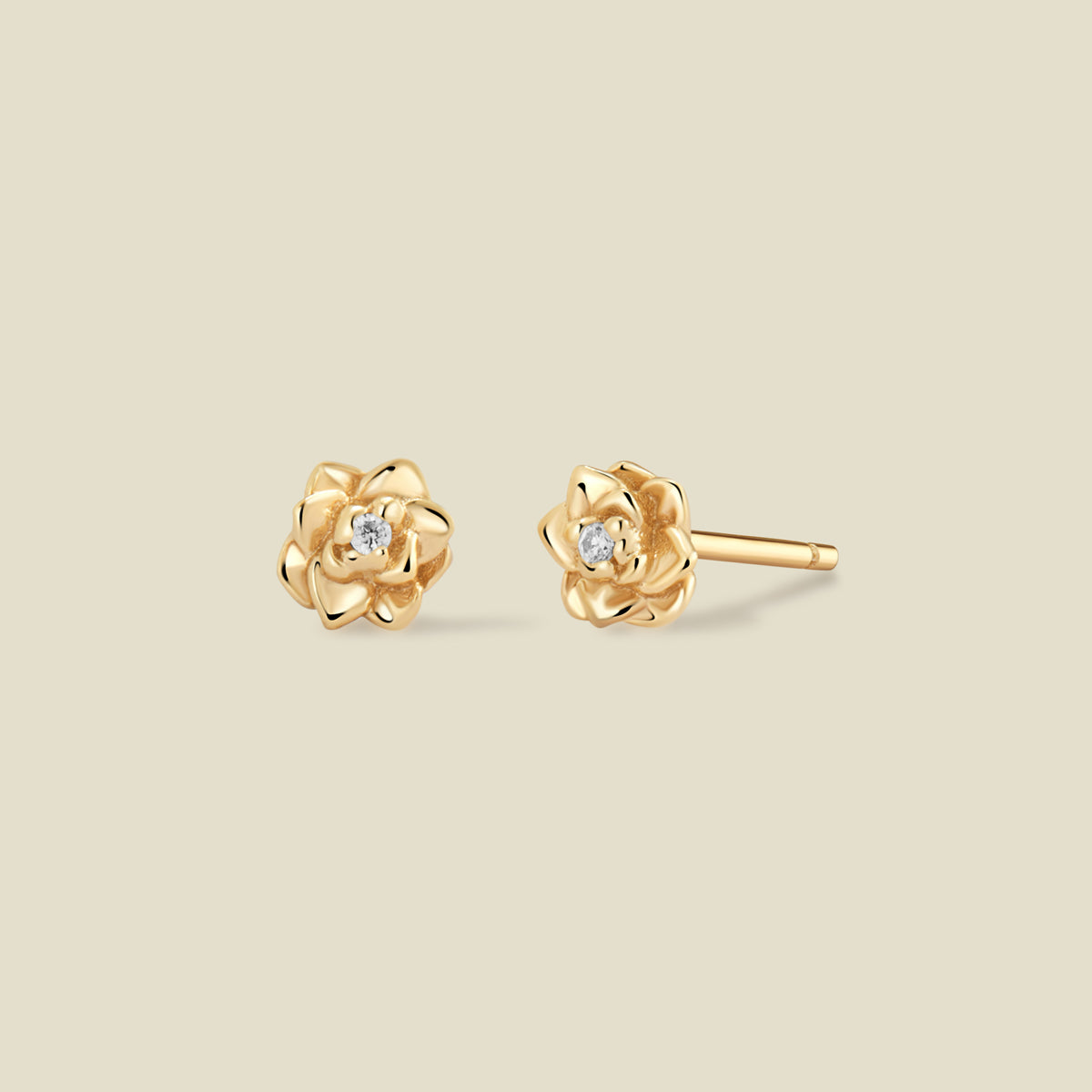 A pair of gold lotus flower stud earrings, each featuring a small round clear gemstone at the center, displayed on a solid light beige background.