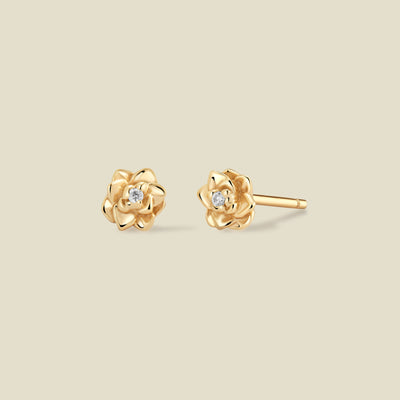 A pair of gold lotus flower stud earrings, each featuring a small round clear gemstone at the center, displayed on a solid light beige background.