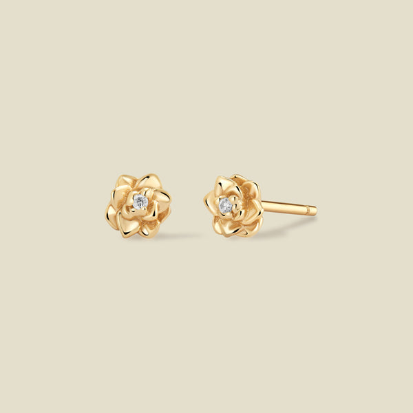 A pair of gold lotus flower stud earrings, each featuring a small round clear gemstone at the center, displayed on a solid light beige background.