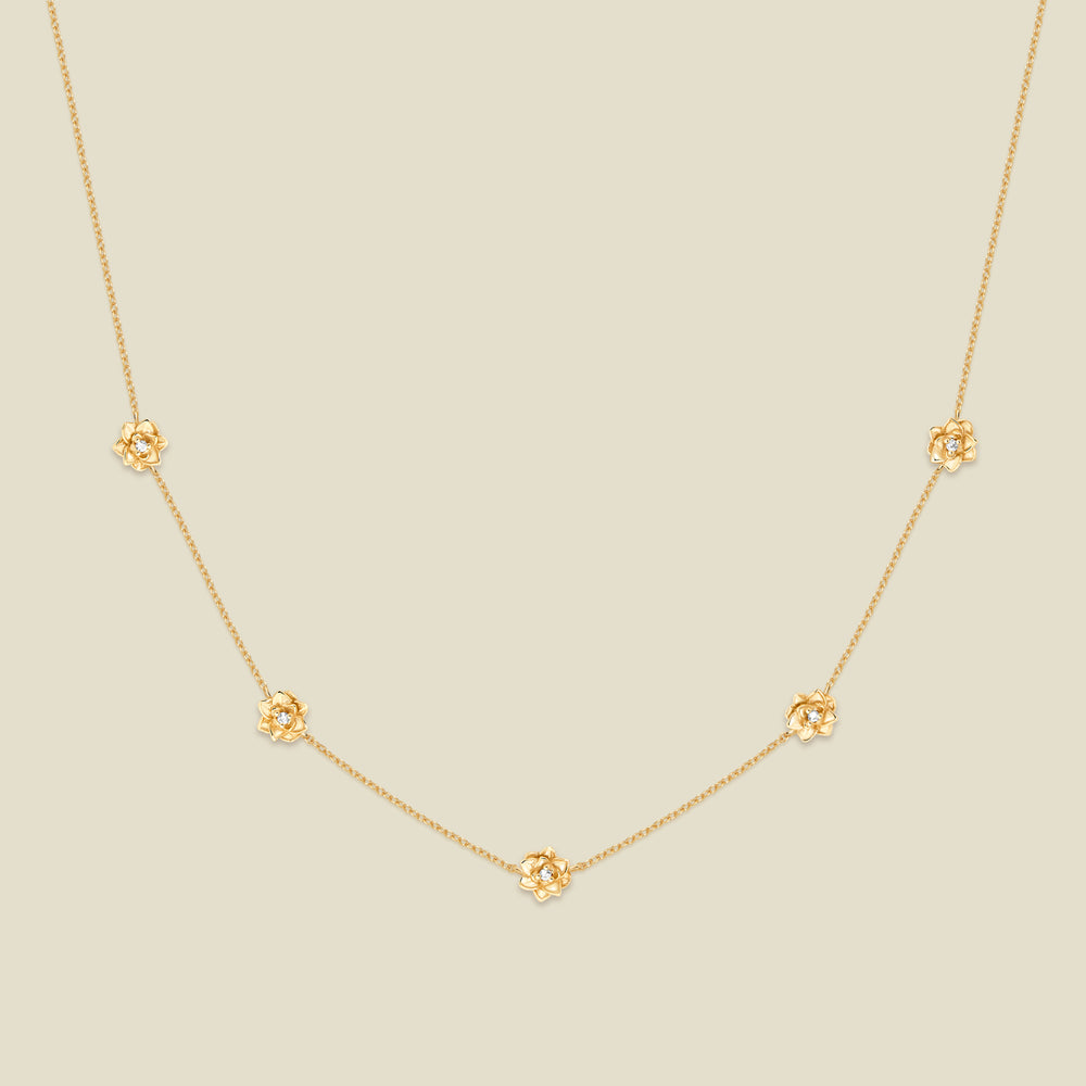 A delicate gold chain necklace featuring five small, evenly spaced textured lotus flower motifs set with tiny clear stones, displayed on a solid light beige background.