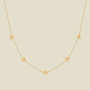 A delicate gold chain necklace featuring five small, evenly spaced textured lotus flower motifs set with tiny clear stones, displayed on a solid light beige background.
