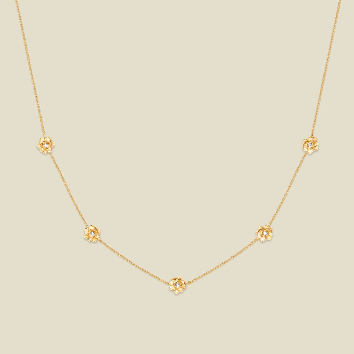 A delicate gold chain necklace featuring five small, evenly spaced textured lotus flower motifs set with tiny clear stones, displayed on a solid light beige background.