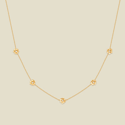 A delicate gold chain necklace featuring five small, evenly spaced textured lotus flower motifs set with tiny clear stones, displayed on a solid light beige background.