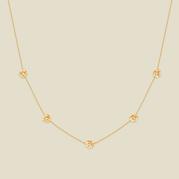 A delicate gold chain necklace featuring five small, evenly spaced textured lotus flower motifs set with tiny clear stones, displayed on a solid light beige background.