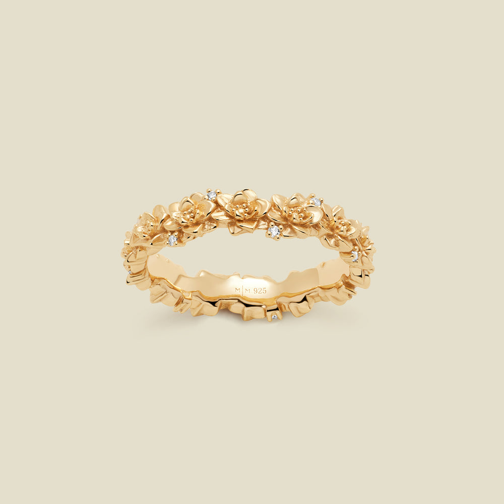 A gold floral-etched ring band with textured lotus flowers and small clear gemstones, shown on a plain warm beige background. 