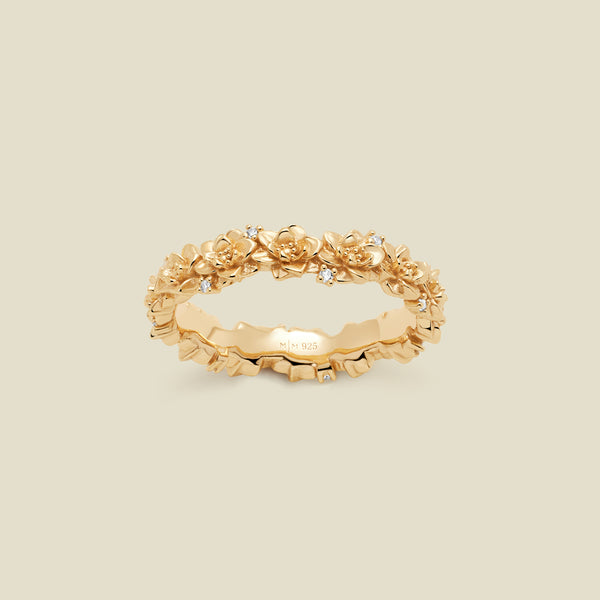 A gold floral-etched ring band with textured lotus flowers and small clear gemstones, shown on a plain warm beige background. 