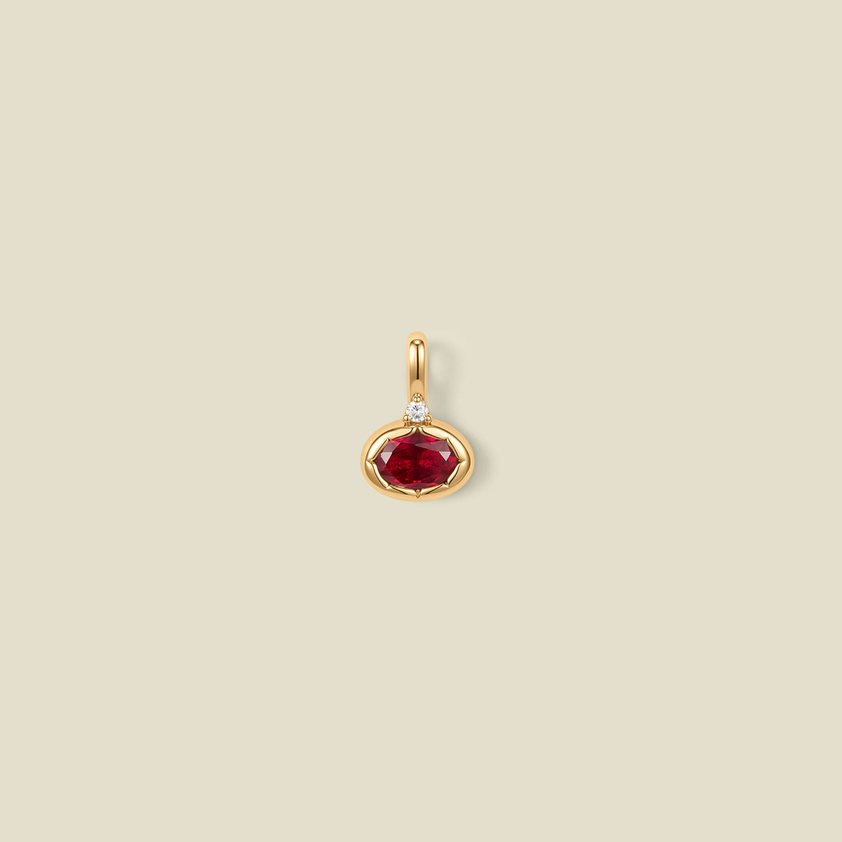 A gold oval pendant charm with a red bezel-set oval gemstone and a clear accent stone on top, set against a solid beige background.