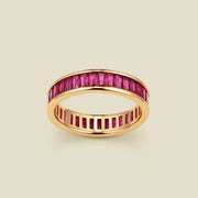 A gold eternity ring featuring a continuous channel setting of baguette-cut red gemstones, shown on a solid light beige background.