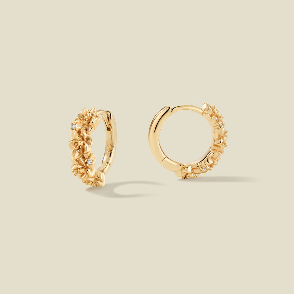 A pair of gold hoop earrings adorned with lotus flowers stacked along the hoop, with scattered small clear accent stones, set against a solid beige background.