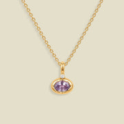 A gold chain necklace with a small oval pendant featuring a bezel set lavender-purple gemstone and a tiny clear accent stone above it, shown on a light beige background.